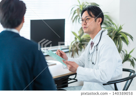 A male doctor, holding a medical chart, listens intently to a patient's story in a consultation room/clinic. (Keywords: health management, regular check-up) 140389058