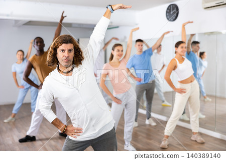 Group of young adult active multiethnic dancers stretching one arm up during learning new dance exercise movement in gym 140389140