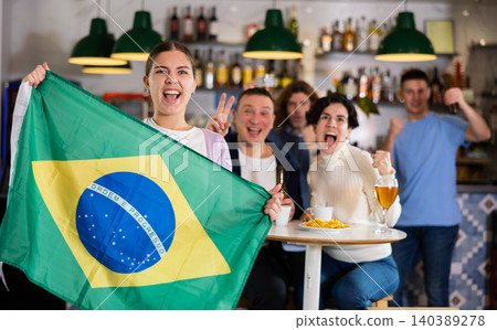 Group of friends, sports supporters cheering for favorite team with flag of Brazil while watching match on TV in bar Group of friends, sports supporters cheering for favorite team with flag of Brazil while watching match on TV in bar 140389278