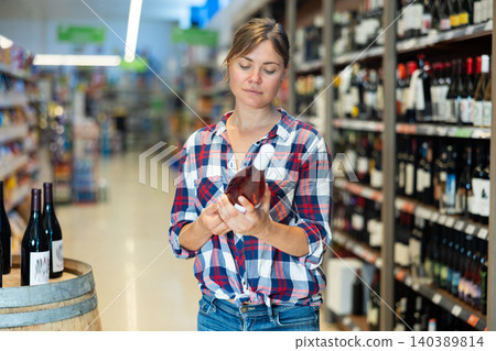 Young woman choosing bottle of wine while shopping in supermarket Young woman choosing bottle of wine while shopping in supermarket 140389814