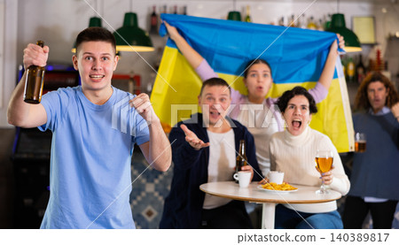 Happy friends of different ages drinking beer and celebrating the victory of the Ukrainian team in night bar Happy friends of different ages drinking beer and celebrating the victory of the Ukrainian team in night bar 140389817