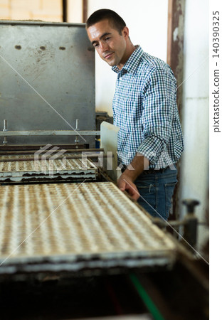 Farmer man working with automatic filling and seeding line for growing containerized seedlings of decorative plants in greenhouse Farmer man working with automatic filling and seeding line for growing containerized seedlings of decorative plants in greenhouse 140390325