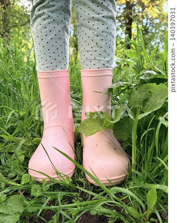 Close-up of a little girl's legs in pink rubber boots standing in wet green grass. Childhood joys Close-up of a little girl's legs in pink rubber boots standing in wet green grass. Childhood joys 140397031