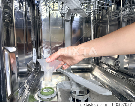 A woman's hand in an open dishwasher holds a plastic funnel. Replenishing the dishwasher salt supply ensures proper water softening and cleaning efficiency. A woman's hand in an open dishwasher holds a plastic funnel. Replenishing the dishwasher salt supply ensures proper water softening and cleaning efficiency. 140397037