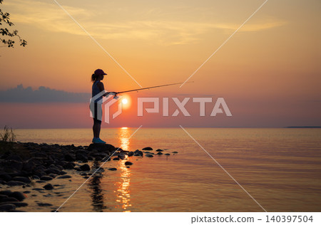 Silhouette of woman fishing on rocky lakeshore at sunset. 140397504
