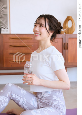 一位微笑的年輕女子在家中做瑜珈休息時喝水。 一位微笑的年輕女子在家中做瑜珈休息時喝水。 140400408