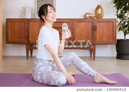 一位微笑的年輕女子在家中做瑜珈休息時喝水。 一位微笑的年輕女子在家中做瑜珈休息時喝水。 140400418