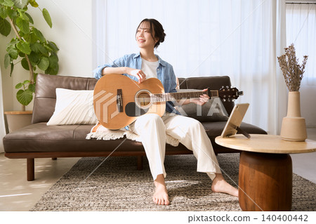 A smiling young woman playing an acoustic guitar on the living room sofa. A smiling young woman playing an acoustic guitar on the living room sofa. 140400442