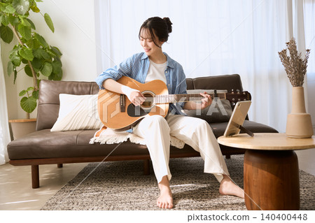 一位面帶微笑的年輕女子在客廳沙發上彈奏原聲吉他。 一位面帶微笑的年輕女子在客廳沙發上彈奏原聲吉他。 140400448