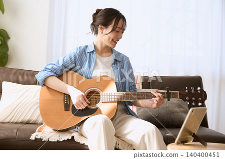 一位面帶微笑的年輕女子在客廳沙發上彈奏原聲吉他。 一位面帶微笑的年輕女子在客廳沙發上彈奏原聲吉他。 140400451