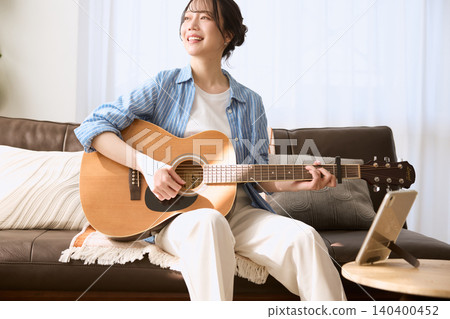 一位面帶微笑的年輕女子在客廳沙發上彈奏原聲吉他。 一位面帶微笑的年輕女子在客廳沙發上彈奏原聲吉他。 140400452