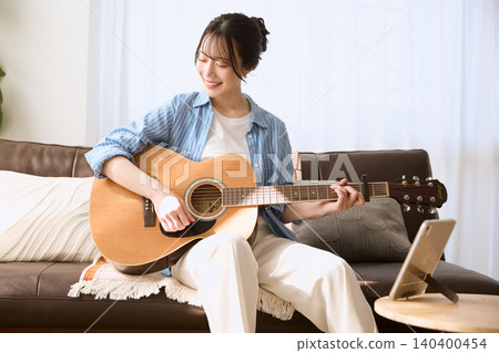 A smiling young woman playing an acoustic guitar on the living room sofa. A smiling young woman playing an acoustic guitar on the living room sofa. 140400454