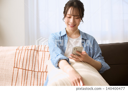一位年輕女子坐在沙發上，看著她的智慧型手機，面帶微笑。 140400477