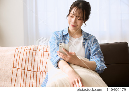 A young woman smiles while looking at her smartphone on the living room sofa. A young woman smiles while looking at her smartphone on the living room sofa. 140400479