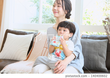 A toddler sits on his mother's lap on the living room sofa and drinks a beverage from a pouch. 140400602