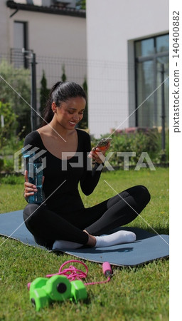 Vertical video. Young sport woman resting and using smartphone, drink water after workout in backyard. Sports female doing outdoor training. Vertical video. Young sport woman resting and using smartphone, drink water after workout in backyard. Sports female doing outdoor training. 140400882