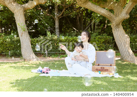 A mother and her young son enjoying a picnic in the park, blowing soap bubbles. 140400954