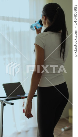 Vertical video. Athletic woman in sportswear running and drink water on treadmill during home workout. Back view. Vertical video. Athletic woman in sportswear running and drink water on treadmill during home workout. Back view. 140400985