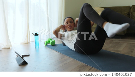 Young woman makes press workout on the mat while watching training video on tablet at home. Woman in sportswear doing exercise raising legs and pumping the press. Fitness training for burning fat 140401027
