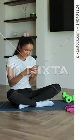Vertical Video. Portrait of a sporty woman resting at home after training and using her smartphone. Beautiful healthy sportive girl athlete resting after intense hard training. 140401124