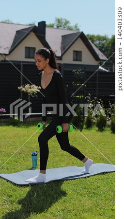 Vertical video. Athletic young woman in sportswear training on the lawn of the house, performing squats with dumbbells in her hands, enjoying a healthy lifestyle. Athletic woman doing training Vertical video. Athletic young woman in sportswear training on the lawn of the house, performing squats with dumbbells in her hands, enjoying a healthy lifestyle. Athletic woman doing training 140401309