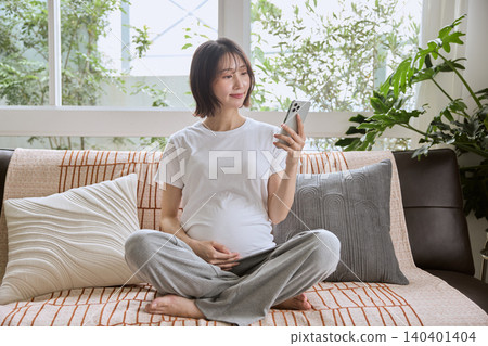 一位孕婦坐在沙發上看著她的智慧型手機。 一位孕婦坐在沙發上看著她的智慧型手機。 140401404