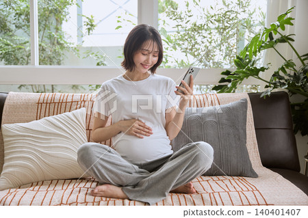 一位孕婦坐在沙發上,看著她的智慧型手機,並將手放在肚子上。 一位孕婦坐在沙發上,看著她的智慧型手機,並將手放在肚子上。 140401407