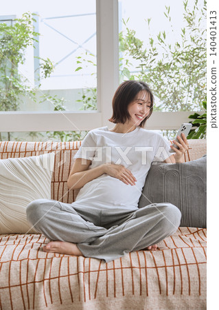 一位年輕的孕婦放鬆地躺在沙發上,看著她的智慧型手機,笑著。 一位年輕的孕婦放鬆地躺在沙發上,看著她的智慧型手機,笑著。 140401413