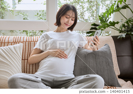 一位孕婦坐在沙發上，看著她的智慧型手機，並將手放在肚子上。 140401415