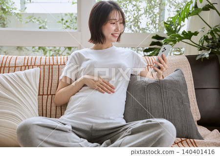 A pregnant woman sits on a sofa, looking at her smartphone and placing her hand on her stomach. 140401416