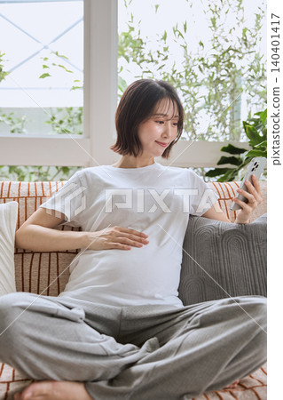 一位孕婦放鬆地躺在客廳沙發上,看著她的智慧型手機。 一位孕婦放鬆地躺在客廳沙發上,看著她的智慧型手機。 140401417