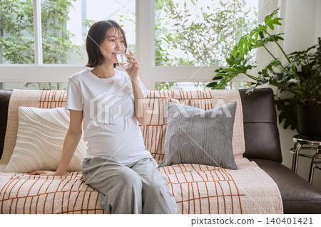 A pregnant woman drinking a glass of water on the living room sofa. A pregnant woman drinking a glass of water on the living room sofa. 140401421