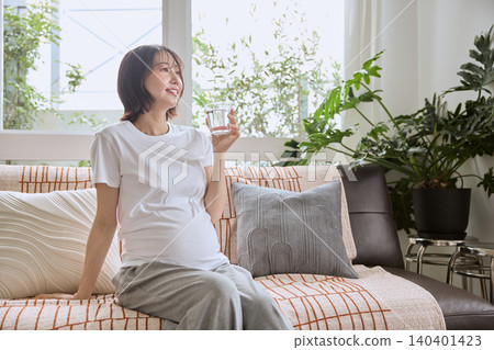 A pregnant woman drinking a glass of water on the living room sofa. 140401423