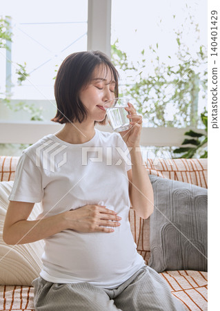 一位孕婦坐在沙發上,一邊喝水一邊撫摸著肚子。 一位孕婦坐在沙發上,一邊喝水一邊撫摸著肚子。 140401429