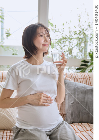 A pregnant woman sits on a sofa, holding her stomach and drinking water. 140401430