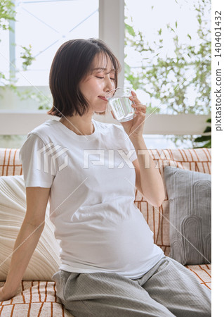 A young pregnant woman in a white T-shirt sits on a sofa and drinks a glass of water. A young pregnant woman in a white T-shirt sits on a sofa and drinks a glass of water. 140401432