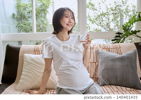 一位孕婦坐在客廳沙發上喝水。 140401435