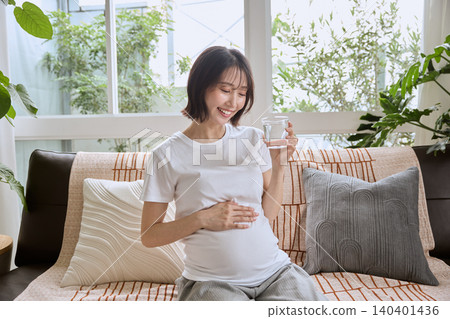 一位孕婦在客廳沙發上喝水,臉上帶著微笑。 一位孕婦在客廳沙發上喝水,臉上帶著微笑。 140401436