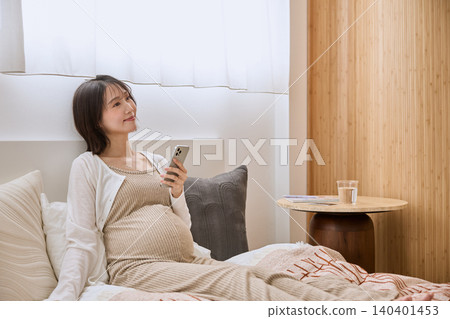 一位孕婦躺在床上,一邊看著手機一邊微笑。 一位孕婦躺在床上,一邊看著手機一邊微笑。 140401453