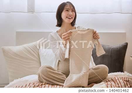 一位年輕女子麵帶微笑,手裡拿著嬰兒衣服放在床上。 一位年輕女子麵帶微笑,手裡拿著嬰兒衣服放在床上。 140401462