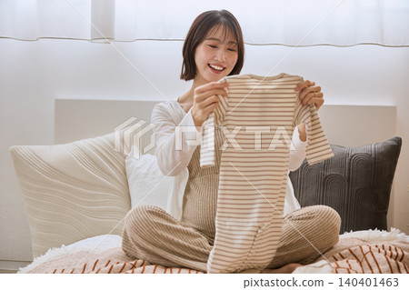 一位孕婦微笑著在床上鋪開嬰兒衣服。 一位孕婦微笑著在床上鋪開嬰兒衣服。 140401463