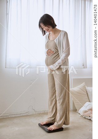 A pregnant woman smiles as she steps on a scale in her bedroom and places her hand on her stomach. 140401478