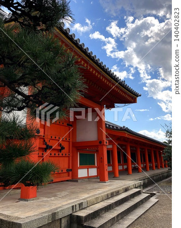 A vermilion-painted traditional Japanese building and pine tree stand beneath a blue sky and clouds. A vermilion-painted traditional Japanese building and pine tree stand beneath a blue sky and clouds. 140402823