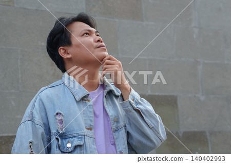 Thoughtful Young Asian Man Holding Chin and Thinking Against Stone Wall Thoughtful Young Asian Man Holding Chin and Thinking Against Stone Wall 140402993