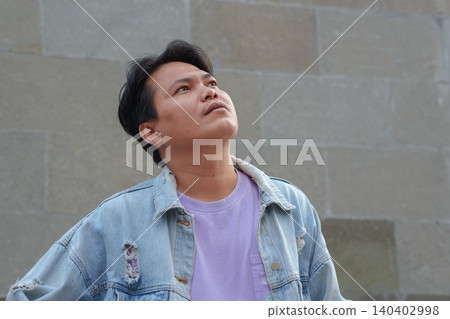 Thoughtful Young Asian Man Wearing Denim Jacket Looking Up Against Stone Wall 140402998