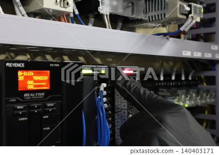 A close-up of an industrial control panel where a person operating a Keyence PLC module with a hand wearing a black glove. 140403171