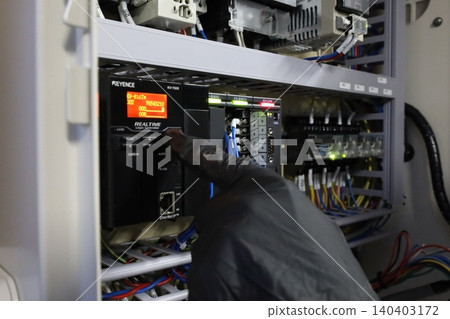 An engineer's hand inspecting a Keyence PLC module inside an industrial control cabinet. 140403172
