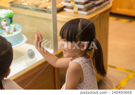 Excited little Asian girl looking at beautiful cake through glass display in a bakery shop, childhood birthday wishes. 140403608