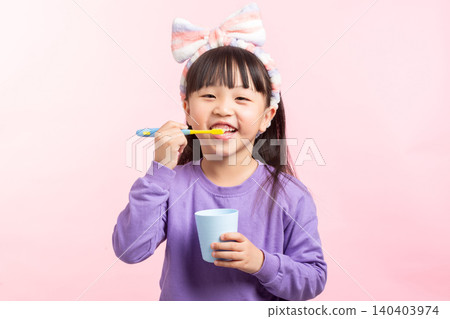 A young Asian elementary girl with long dark hair, bangs, and a striped headband, wearing a purple sweatshirt, happily brushing her teeth while holding a blue cup against a solid light pink studio 140403974