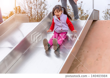 a cheerful girl sliding down a steel slide in the playground with open arms. a cheerful girl sliding down a steel slide in the playground with open arms. 140403984